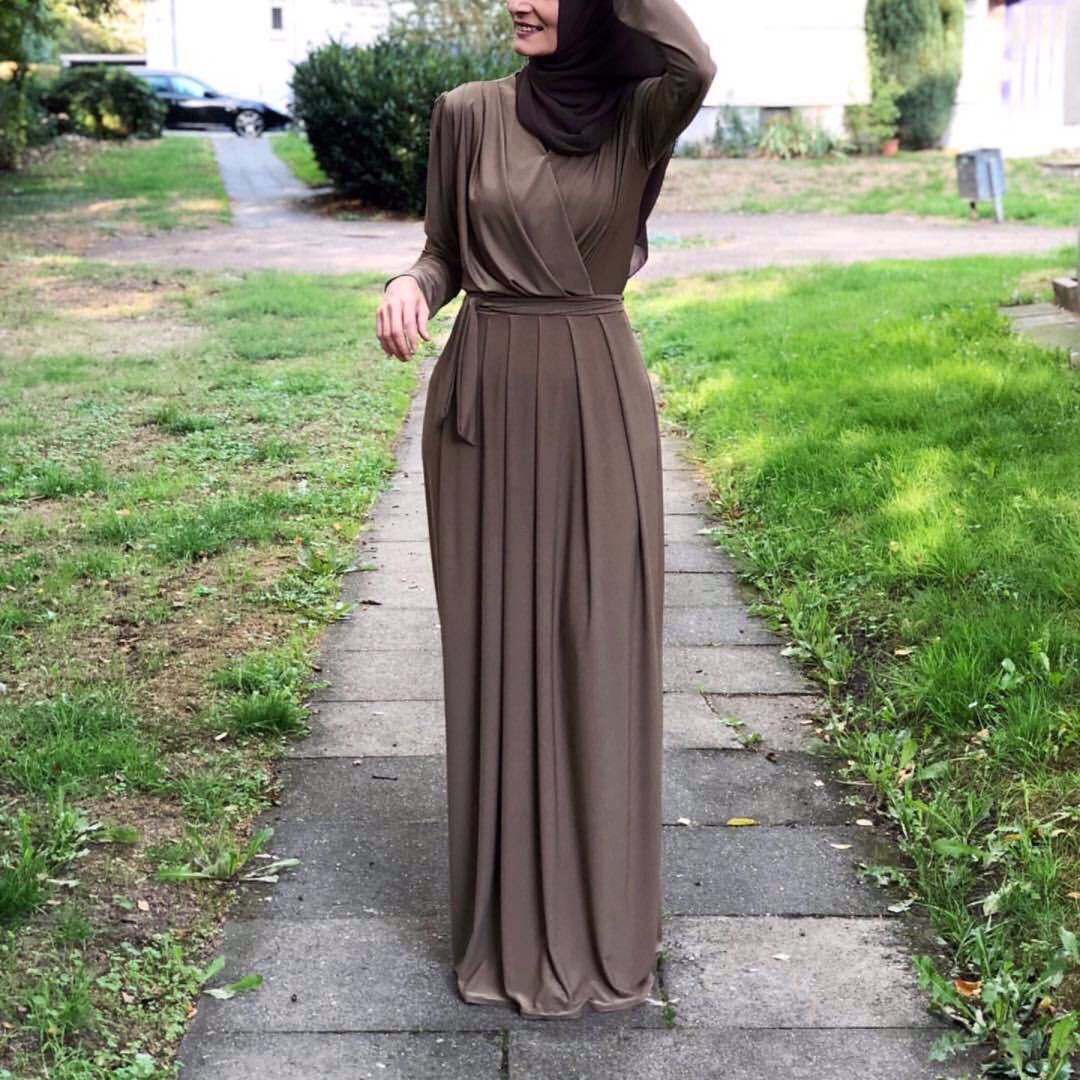 Woman wearing a brown pleated maxi skirt with a V-neck design, walking on a pathway surrounded by greenery.