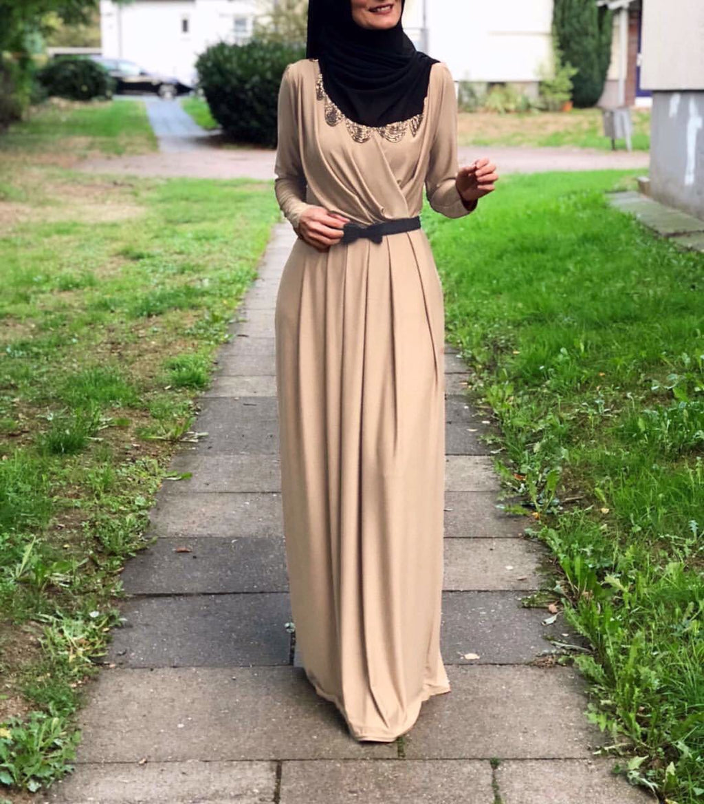Woman in a beige V-neck tie pleated maxi skirt with a black belt, walking on a sidewalk in a green area.
