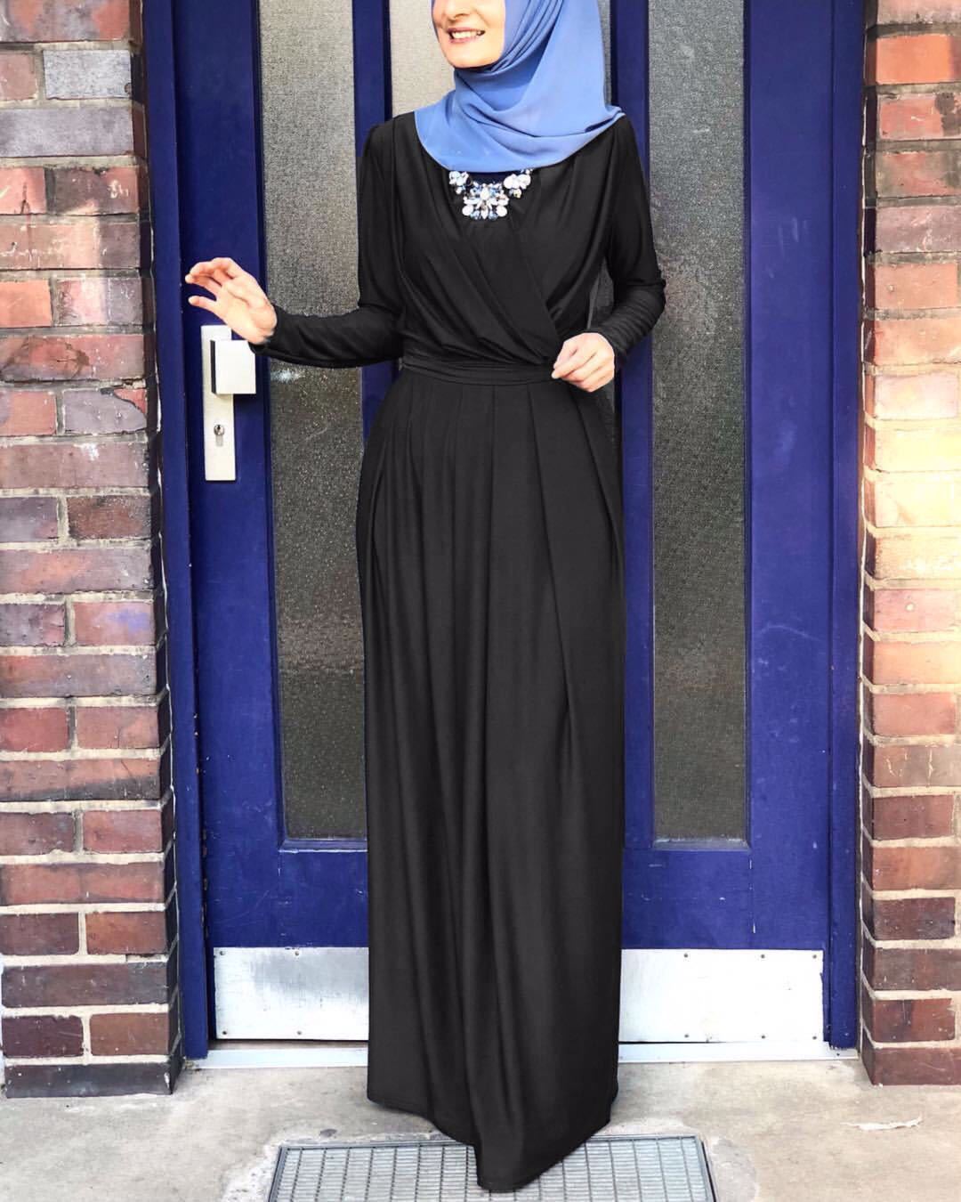 Elegant woman in a V-neck black pleated maxi skirt with a blue hijab and statement necklace, standing by a blue door.
